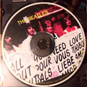 May include: A white vinyl record with a black and white photo of The Beatles on the cover. The text "All You Need Is Love" is written in a circle around the photo. The text is in multiple languages, including English, French, German, and Russian.