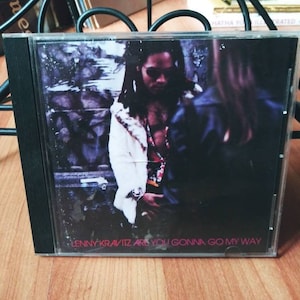 May include: A CD case with a black background and a photo of Lenny Kravitz on the front. The text "Lenny Kravitz Are You Gonna Go My Way" is printed in pink on the bottom of the case.