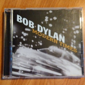 May include: A black CD case with a blurry image of a car on the front. The text "BOB DYLAN MODERN TIMES" is printed in gold on the front of the case.