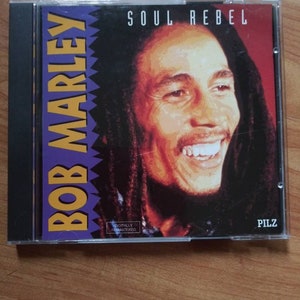 May include: A compact disc with a purple, yellow, and black design. The album cover features a photo of Bob Marley with the text "Soul Rebel" and "Bob Marley" in large, bold letters. The text "Digitally Remastered" and "Pilz" are also visible on the CD.