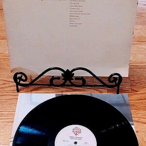 May include: A black vinyl record with a white label that reads "James Taylor's Greatest Hits". The record is on a white surface with a black metal record stand.