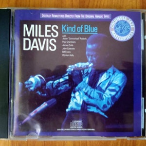 May include: A CD case with a blue and black cover featuring a photo of Miles Davis playing a trumpet. The title of the album is "Kind of Blue" and the text "Columbia Jazz Masterpieces" is printed on the cover.