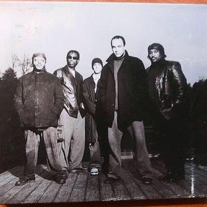 May include: A group of five people stand on a wooden deck, posing for a photograph. They are all wearing dark-colored jackets and coats. The image is in black and white.