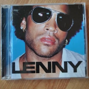 May include: A CD case with a photo of Lenny Kravitz on the front. He is wearing sunglasses and a white shirt. The text "LENNY" is printed in black on the front of the case.