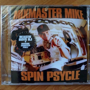 May include: A CD case with a black and orange design. The front features a photo of a man wearing a black baseball cap and a white shirt. The text "Mixmaster Mike" is in large orange letters at the top of the case. The text "Spin Psycle" is in large orange letters at the bottom of the case. A smaller circle with the text "Beastie Boys DJ" is in the upper left corner of the case.