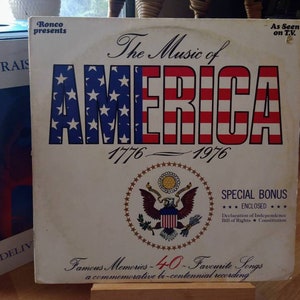 May include: A white vinyl record album cover with a red, white, and blue American flag design. The title "The Music of America 1776-1976" is printed in black cursive font. The album cover features a special bonus enclosed: Declaration of Independence, Bill of Rights, Constitution.  The album cover also includes the text "Famous Memories - 40 - Favourite Songs a commemorative bi-centennial recording".