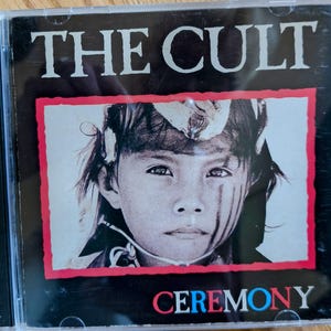 Puede incluir: Una caja de CD con el álbum "Ceremony" de The Cult. La portada tiene un fondo negro con el nombre de la banda en blanco. La imagen central muestra a un niño con pintura facial. La palabra "CEREMONY" está en rojo, blanco y azul.