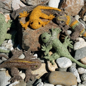 May include: A collection of five handcrafted felt lizard figures in shades of green, brown, and yellow, displayed on a rocky surface. The lizards feature intricate details and are positioned in various poses, highlighting their individual characteristics.