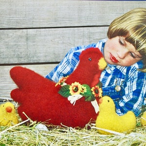 May include: A red knitted hen with a floral wreath around its neck and four yellow knitted chicks in a nest of hay. A young child with blonde hair is lying on the hay next to the hen and chicks.