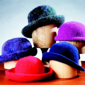 May include: Four felt hats in different colors, including green, purple, blue, and red, are displayed on wooden hat forms. The hats are all the same style, with a round crown and a wide brim.