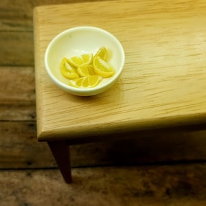 May include: A small, off-white ceramic bowl filled with miniature yellow lemon wedges. The bowl sits on a light-colored wooden table with a single visible leg. The lemons are bright yellow with white details, suggesting a food-themed item.
