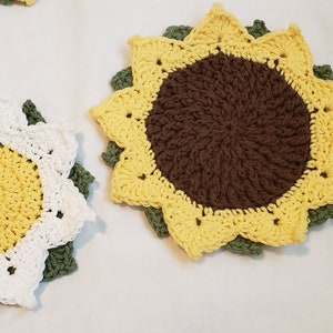 May include: Crocheted sunflower coasters in yellow, brown, white, and green. The coasters are handmade with a detailed petal design and a central circle. The coasters are placed on a white surface.