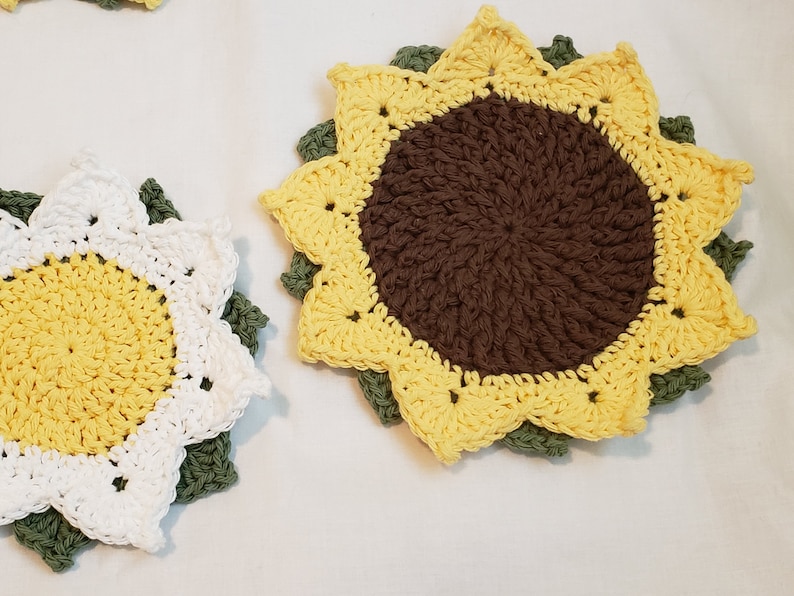 May include: Crocheted sunflower coasters in yellow, brown, white, and green. The coasters are handmade with a detailed petal design and a central circle. The coasters are placed on a white surface.