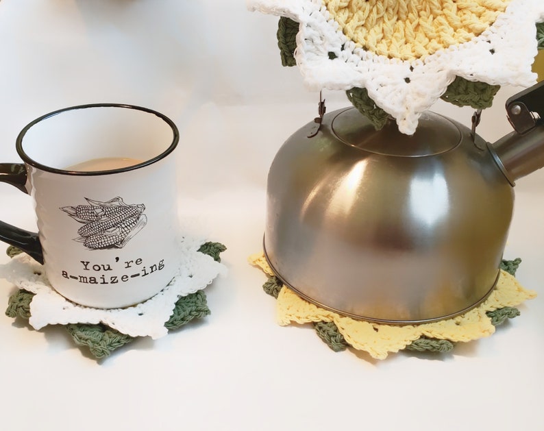 May include: A white enamel mug with a black rim and handle, featuring a corn graphic and the text "You're a-maize-ing." The mug sits on a white and green crocheted coaster. A stainless steel kettle rests on a yellow, white, and green crocheted coaster.