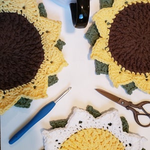 May include: Crocheted sunflower coasters in yellow, brown, and green. A blue crochet hook and bronze scissors are also present. A black mug is in the background. The coasters are handmade and have a detailed, textured design.