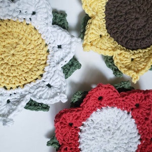 May include: Close-up of four handmade crochet coasters in the shape of flowers. The coasters feature yellow, white, and red petals with green leaves. The center of one coaster is brown. The coasters are on a white surface.
