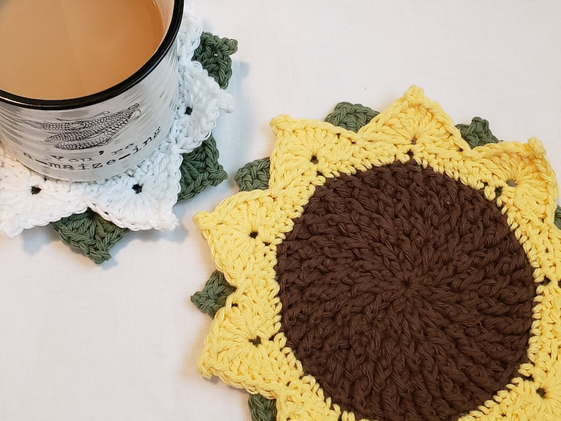 May include: Two handmade crochet coasters in the shape of sunflowers. One coaster is under a white mug with a beverage. The coasters feature yellow, brown, white, and green colors. The mug has the text "You're amazing".