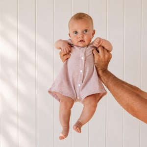 May include: A light dusty pink baby romper featuring a sleeveless design, a round neckline, and a vertical row of four small white buttons down the centre front. The garment has a loose, flared fit around the legs and is made of a textured fabric.