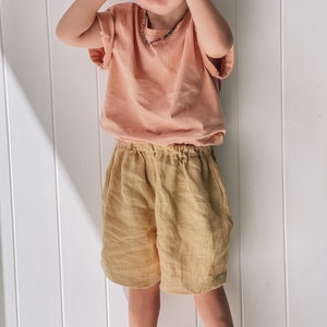 May include: A young child wearing a peach-colored t-shirt and beige linen shorts. The child is standing in front of a white wall with vertical panels.