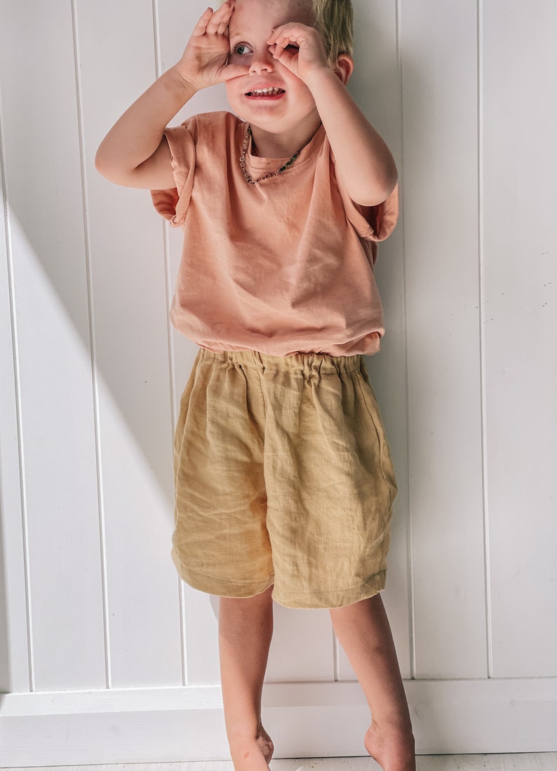 May include: A young child wearing a peach-colored t-shirt and beige linen shorts. The child is standing in front of a white wall with vertical panels.