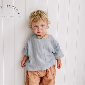 May include: A young child wearing a light blue linen top with a gold necklace and brown pants. The child is standing in front of a white wall with vertical panels. The text "TINY DESIGN SEWING PATTERNS" is visible in the top left corner of the image.