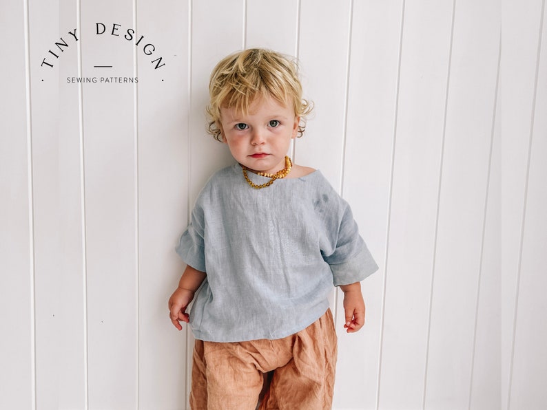 May include: A young child wearing a light blue linen top with a gold necklace and brown pants. The child is standing in front of a white wall with vertical panels. The text "TINY DESIGN SEWING PATTERNS" is visible in the top left corner of the image.