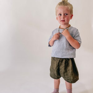 May include: A young boy with blonde hair in a bun is wearing a light blue linen shirt, olive green linen shorts, and pink socks. He is wearing a beaded necklace with amber and green beads.