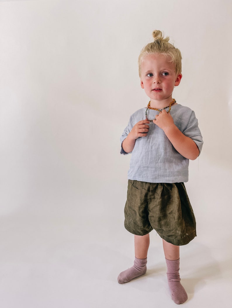 May include: A young boy with blonde hair in a bun is wearing a light blue linen shirt, olive green linen shorts, and pink socks. He is wearing a beaded necklace with amber and green beads.
