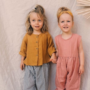May include: Two young children in children's clothing. One wears a mustard button-down shirt with rolled sleeves and blue and white striped trousers. The other wears a pink jumpsuit and a yellow headband. The clothing appears to be made of linen.