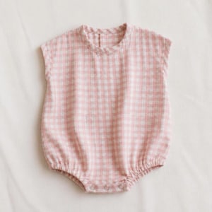 Pode incluir: Um macacão de bebé rosa e branco com padrão xadrez. A peça única tem decote redondo e mangas curtas. Apresenta cintura e aberturas das pernas franzidas e um fecho de botão. O tecido é macio e leve.