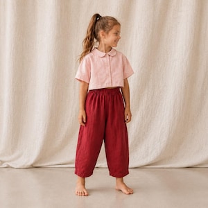 May include: A short-sleeved, light pink button-down shirt with a rounded collar paired with loose-fitting, dark red cropped trousers featuring an elastic waistband. The outfit consists of a solid-colored top and matching solid-colored bottoms.
