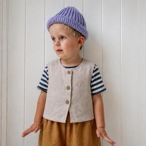 Pode incluir: Um gorro de malha canelada em violeta claro, um colete bege sem mangas com três botões castanhos e uma t-shirt de manga curta com riscas horizontais azul-marinho e brancas, combinada com calças em amarelo mostarda.