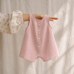 Puede incluir: Un pelele sin mangas en color rosa empolvado, con cuello redondo y una tapeta frontal vertical con cinco pequeños botones blancos. La prenda tiene una silueta sencilla de corte recto y está confeccionada en un tejido con textura.