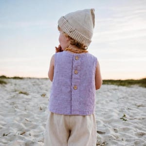 Pode incluir: Uma blusa sem mangas na cor lavanda com uma fileira vertical de três botões de madeira nas costas, combinada com calças bege claro e um gorro de malha bege a condizer. Um colar de contas cor de âmbar é usado à volta do pescoço.
