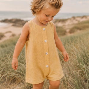 May include: A sleeveless mustard yellow romper featuring a round neckline and a vertical front placket with four light-colored buttons. The garment has a simple, straight-cut silhouette and is constructed from a textured, woven fabric.