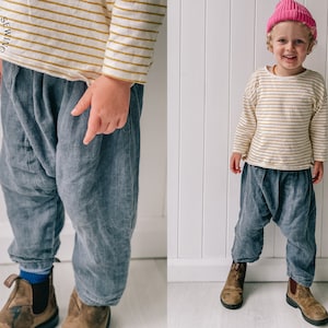 May include: A child wearing a striped long-sleeved shirt and gray pants with a pink beanie. The child is standing in front of a white wall.