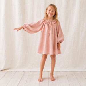 May include: A dusty rose-coloured dress featuring long, voluminous bishop sleeves with gathered cuffs, a round neckline with a delicate tie closure, and a loose, flowing silhouette that falls above the knees in a simple, unadorned design.