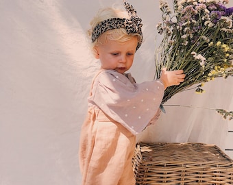May include: A young child wearing a pink and white polka dot shirt and tan pants holds a bouquet of purple and white flowers. The child is wearing a leopard print headband. The child is standing in front of a wicker basket.