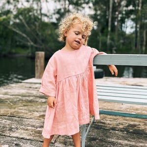 Puede incluir: Una niña pequeña con un vestido de lino rosa está de pie en un muelle de madera, apoyada en un banco azul claro. Tiene el pelo rubio rizado y mira hacia abajo.