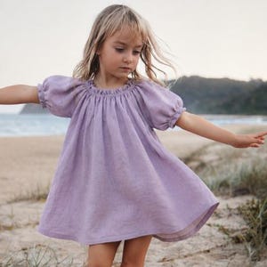 Pode incluir: Um vestido roxo claro com decote redondo franzido e mangas curtas bufantes com punhos elásticos. A peça tem uma silhueta solta e fluida que cai acima do joelho, confeccionada em um tecido com detalhes de textura visíveis.