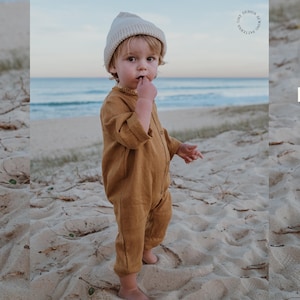 Può includere: Un bambino indossa un pagliaccetto di lino color senape e un berretto color crema su una spiaggia sabbiosa. Il pagliaccetto ha maniche lunghe e bottoni. L'oceano è visibile sullo sfondo. Il testo "TINY DESIGN SEWING PATTERNS" è in un cerchio in alto a destra.