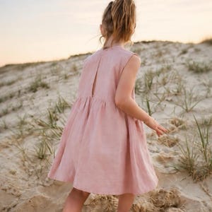 May include: A sleeveless, dusty pink dress featuring a high neckline and a vertical slit opening at the upper back. The garment has a gathered waistline that transitions into a loose, flared skirt that falls above the knees.