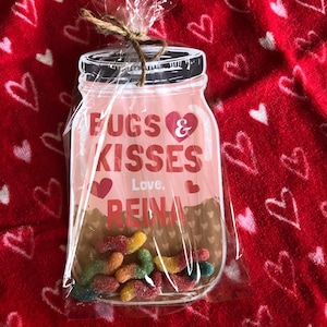 May include: A pink and brown mason jar-shaped card with the text "Bugs & Kisses" and "Love, Reina". The card is filled with colorful gummy worms and is wrapped in clear cellophane.