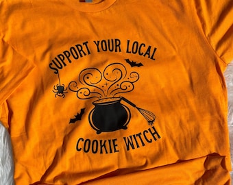 May include: Orange t-shirt with black text that reads "Support Your Local Cookie Witch". The text is accompanied by a black illustration of a cauldron with steam, bats, and a whisk.