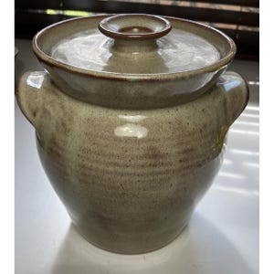 May include: A stoneware crock with a lid, featuring a speckled beige and light brown glaze. The vessel has two side handles and a circular lid with a central knob. The finish is glossy with subtle variations in color throughout the surface.