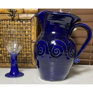 Puede incluir: Una jarra de cerámica azul cobalto con un asa grande y una copa de vino transparente. La jarra presenta diseños de remolinos blancos. La copa de vino tiene una base azul con espirales decorativas.