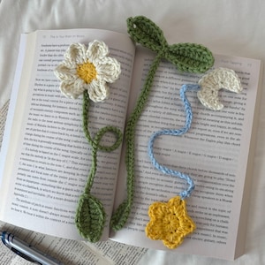 May include: Two handmade crochet bookmarks resting on an open book. One bookmark features a white flower with a yellow center and a green stem with leaves. The other bookmark has a green stem, a light blue section, a white moon, and a yellow star.