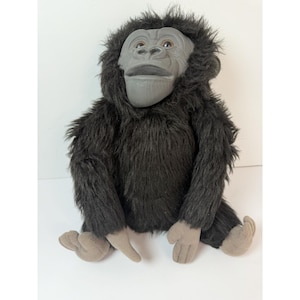 May include: A plush gorilla toy with a gray face and hands, and dark brown fur. The toy is sitting with its legs extended and arms at its sides. The gorilla has a realistic facial expression.