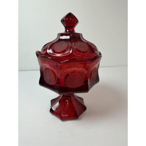 May include: A ruby red glass candy dish with a lid. The dish has a pedestal base and a geometric design with starburst patterns. The lid is topped with a diamond-shaped finial. The dish is likely vintage.