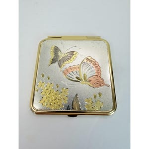 May include: A square compact mirror with a gold-toned metal frame. The lid features a textured silver background decorated with two butterflies in gold and copper tones, alongside a cluster of small gold flowers and a single leaf at the bottom.